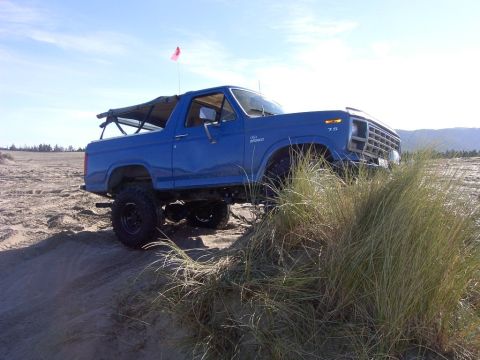 1980 Ford Bronco - Blue 1980 Ford Bronco with soft top parked on a sand dune under a clear sky.