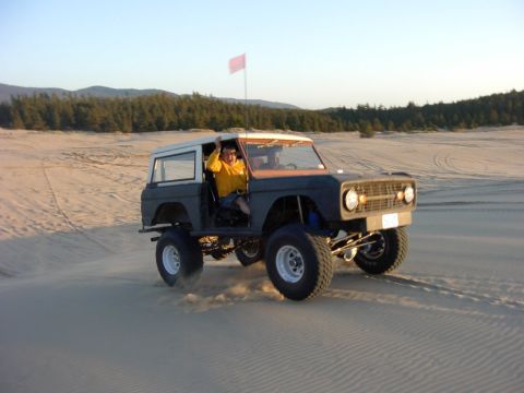 1980 Ford Bronco - A heavily modified Ford Bronco kicks up sand as it drives through a dune landscape under a clear sky.