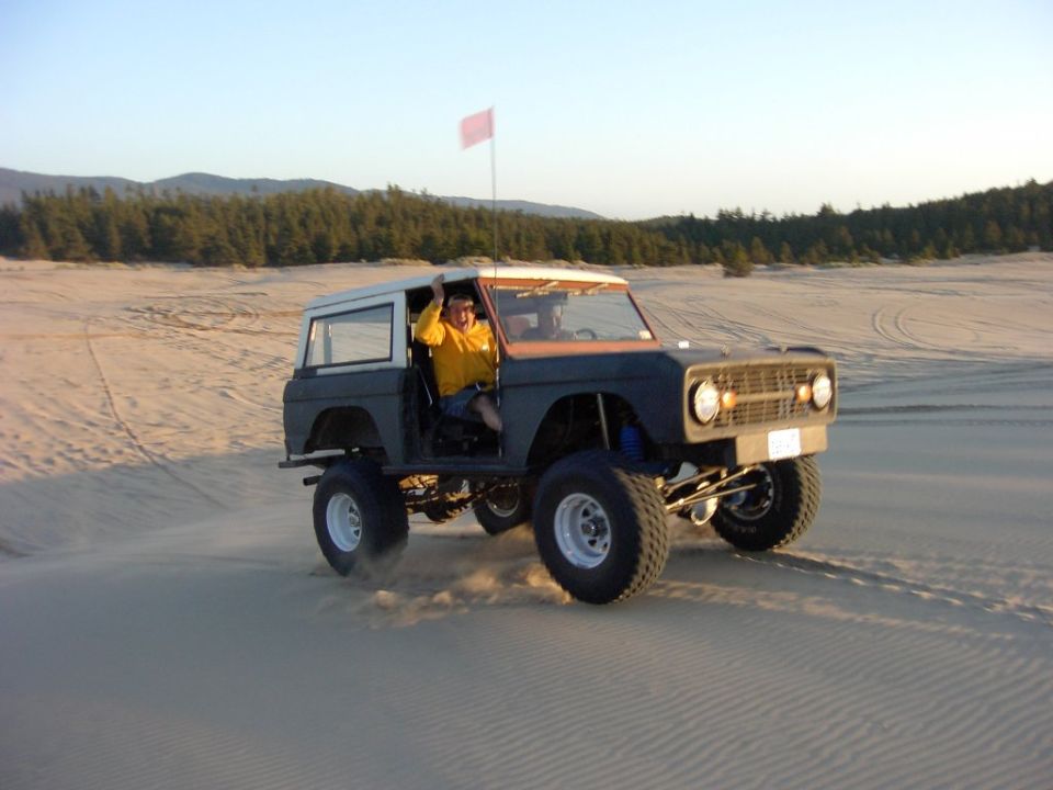 Gray lifted Ford Bronco with white top on sand dunes, driver waving, red flag whip
