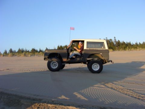 1980 Ford Bronco - A person drives a dark brown and white Ford Bronco across a sandy beach under a clear blue sky.