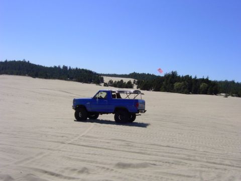 1980 Ford Bronco - A blue 1980 Ford Bronco drives across sandy terrain under a clear blue sky.