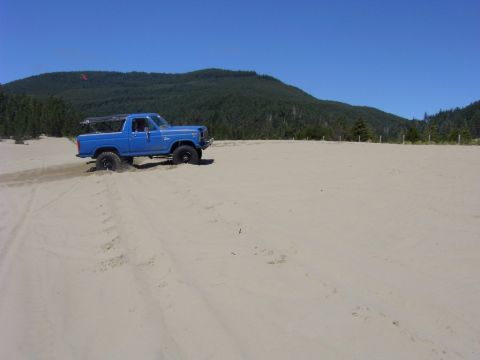 1980 Ford Bronco - A blue pickup truck navigates sandy terrain under a clear blue sky with forested hills in the background.