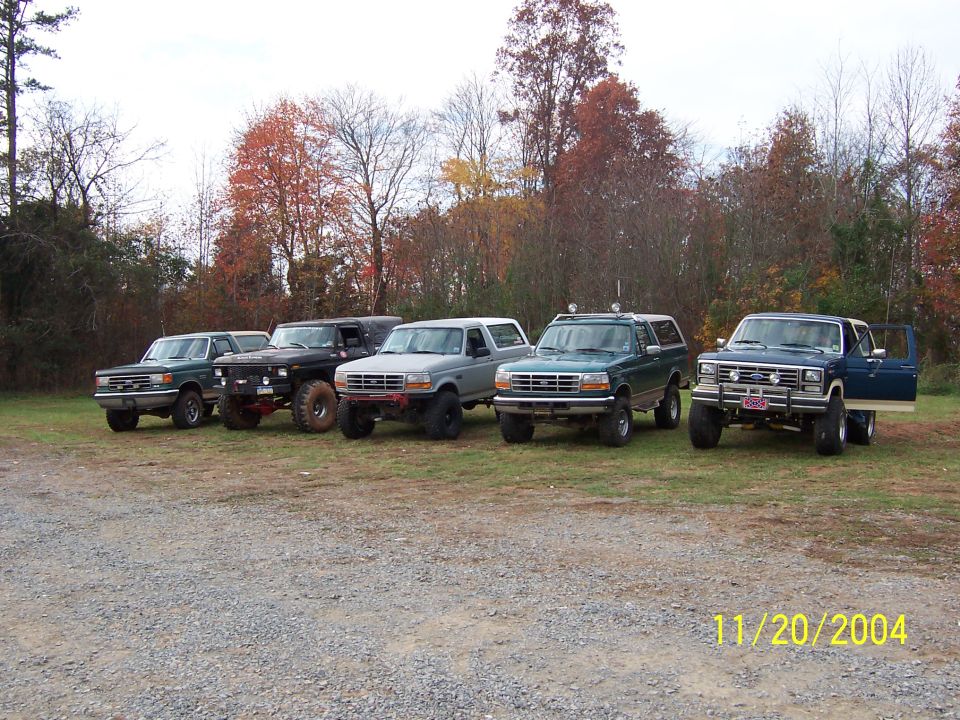 Five Ford Broncos and one F-Series truck parked in a line on a gravel lot with trees in the background