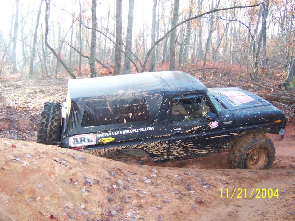 Mud-covered 1978 Ford Bronco tilted in a muddy ditch with trees in the background