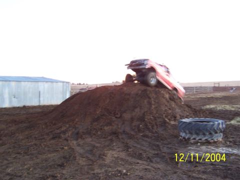 1985 Ford Bronco - A red SUV is seen tipped on its side atop a large dirt mound, with a pair of tires resting on the ground nearby.