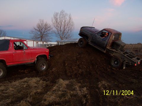 1985 Ford Bronco - Two trucks are shown off-roading on a large dirt hill during dusk.