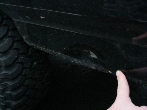 1995 Ford Bronco - Close-up of a black rocker panel and lower door area behind an aggressive mud-terrain tire, with a hand pointing at a dented and creased se…