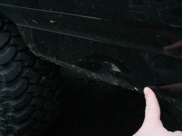 Hand pointing at dent in black rocker panel behind a mud-terrain tire
