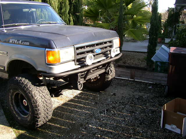 Lifted two-tone blue/silver Ford SUV on mud tires with tube bumper, on gravel driveway