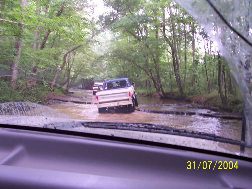 Two trucks driving through a muddy stream in a wooded area. The lead truck is a 1979 Ford F-150