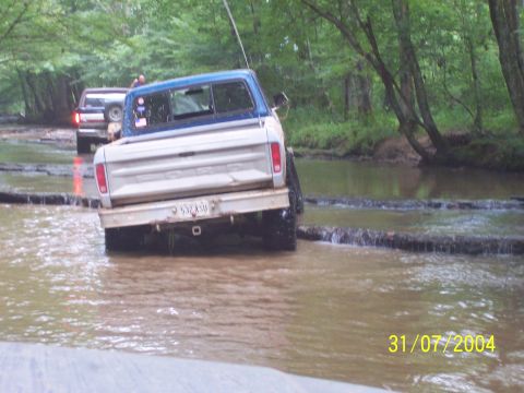 1979 Ford F-150 - Two trucks navigate a water crossing in a wooded area.