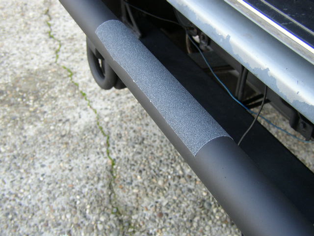 Black tubular side step bar with textured step pad under a vehicle on concrete