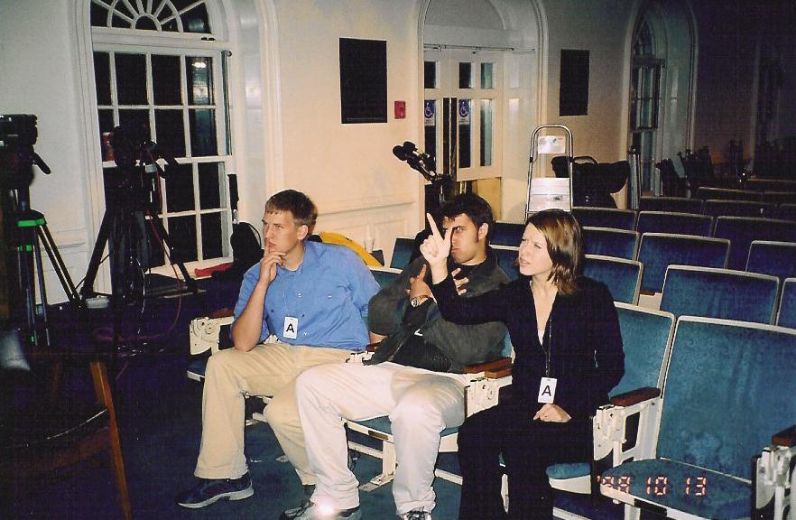 Three people sit in an auditorium with cameras set up. One person points