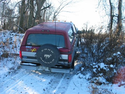1995 Ford Bronco - A red Ford Bronco is parked on a snowy trail surrounded by trees and brush.