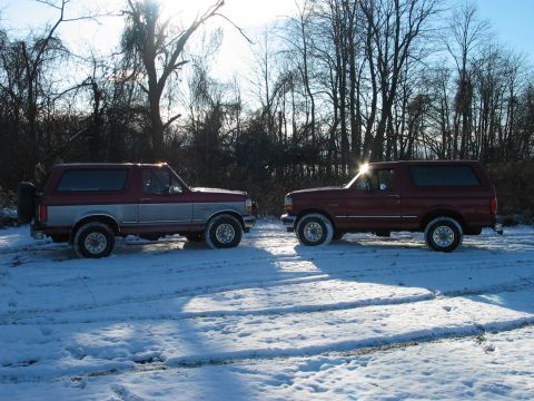 1995 Ford Bronco - Two Ford Broncos sit in a snowy landscape with trees in the background under a bright, hazy sun.