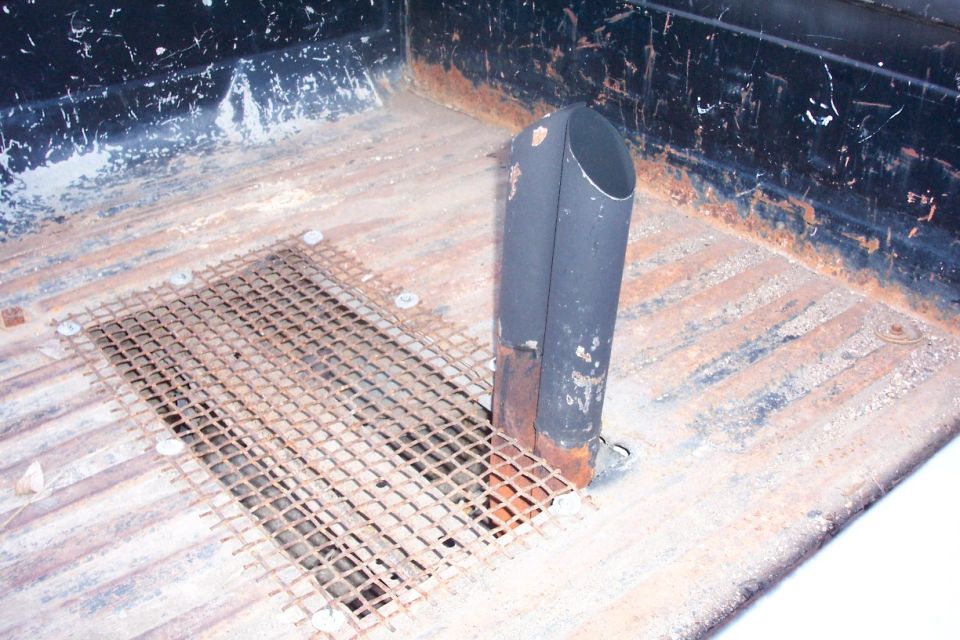Black vertical exhaust stack rising through rusty pickup bed floor with wire mesh patch