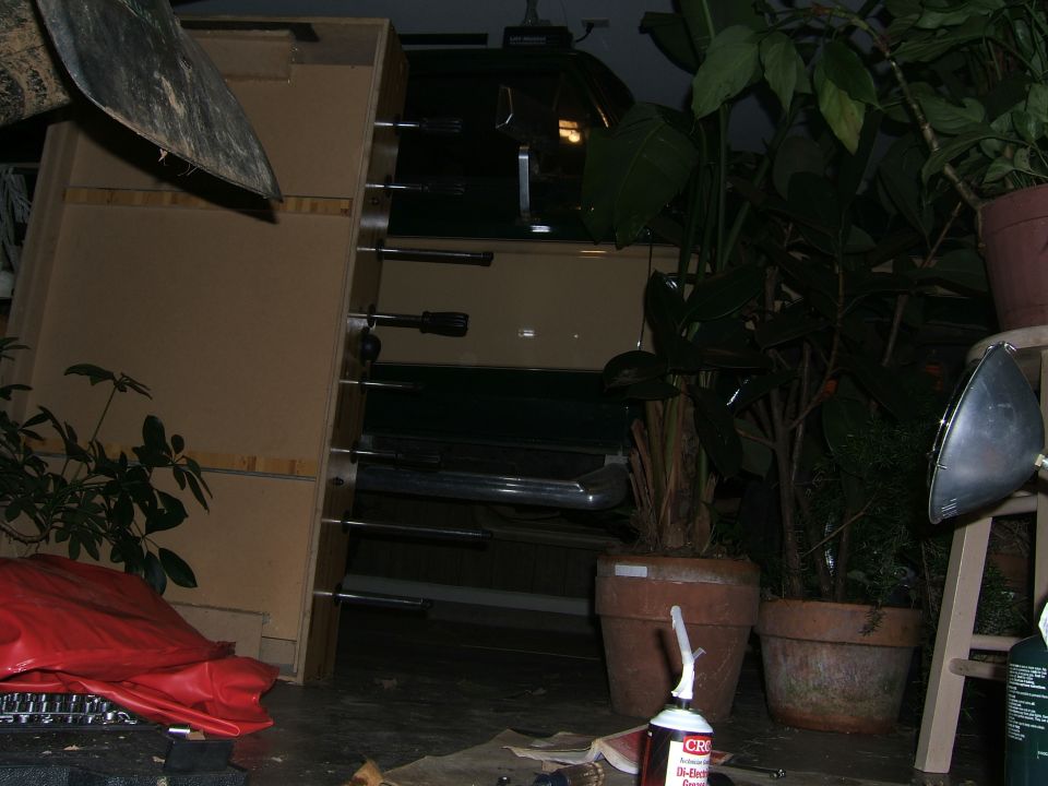 Side view of a tan and dark green vehicle, possibly a 1978 Ford Bronco, partially obscured by plants and storage