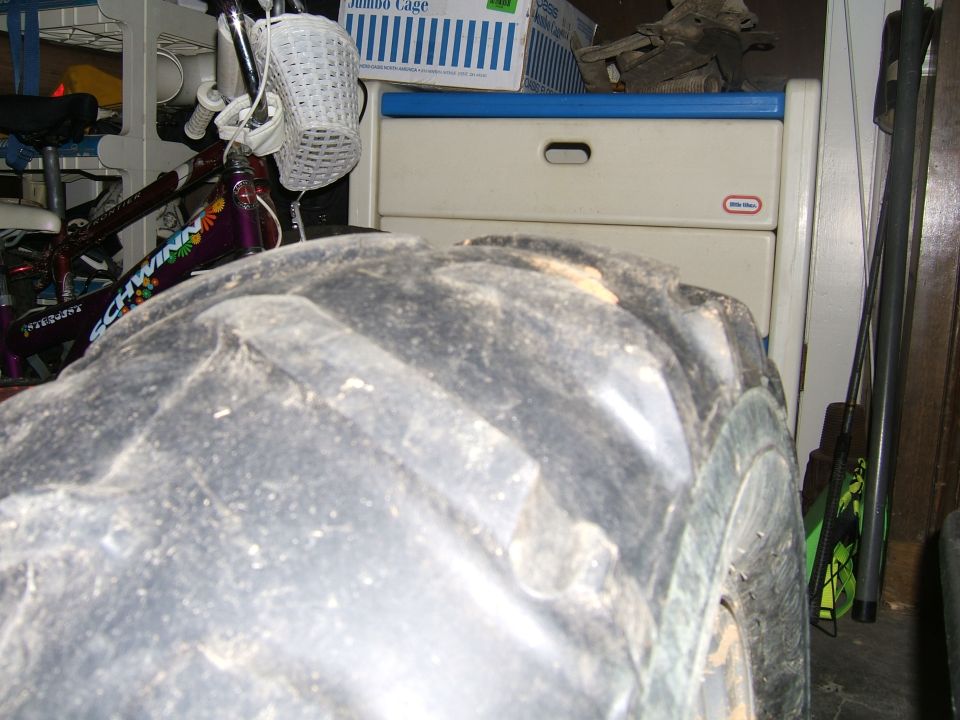 Close-up of a large, knobby off-road tire. A bicycle is visible behind it