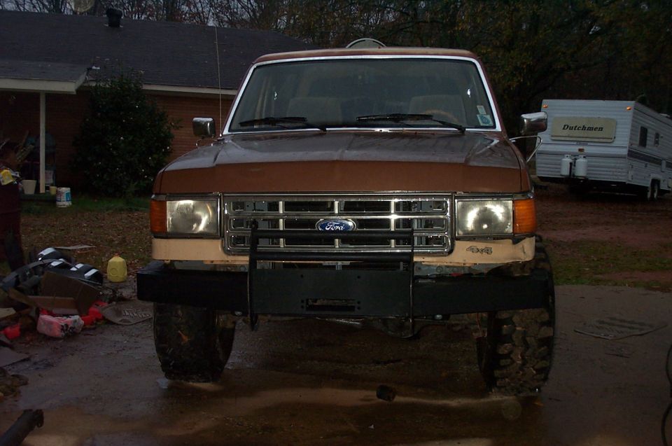 Front view of a brown 1987 Ford Bronco with a new bumper and grille. A trailer is parked behind it