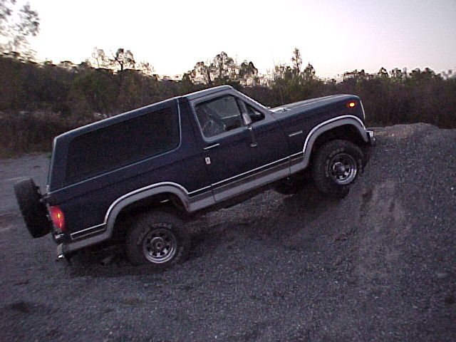 Dark blue two-tone Ford Bronco climbing a gravel mound at dusk, driver side view with rear spare tire