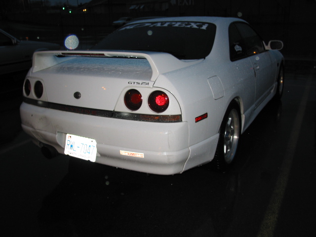 White Nissan Skyline GTS25t rear end in a wet lot at night, Ontario plate and Apexi rear window banner