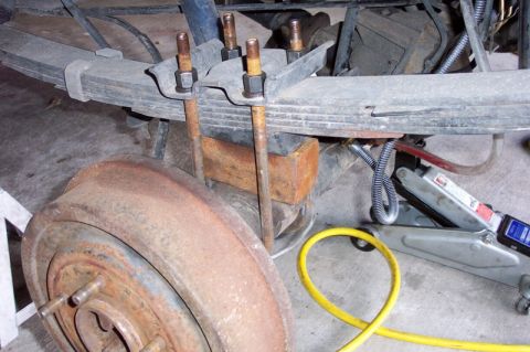 1992 Ford Bronco - Under-vehicle shot of a rear leaf-spring pack clamped to the axle with U-bolts and a rusty lift block between the spring and axle tube. A r…
