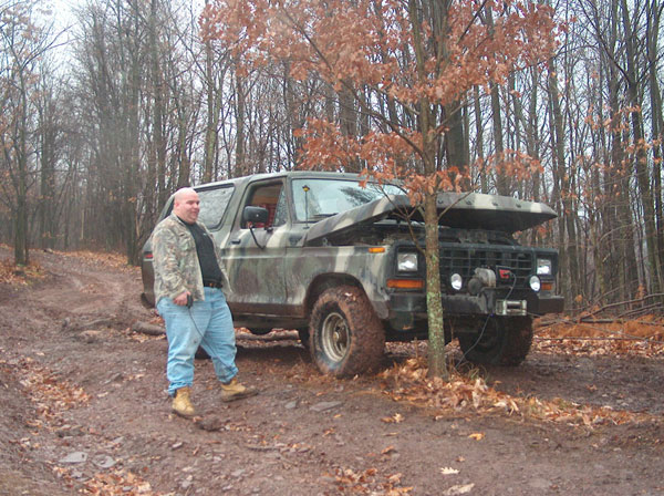 Man in camouflage jacket and jeans stands by a 1978 Ford Bronco with hood open, parked on a muddy forest road