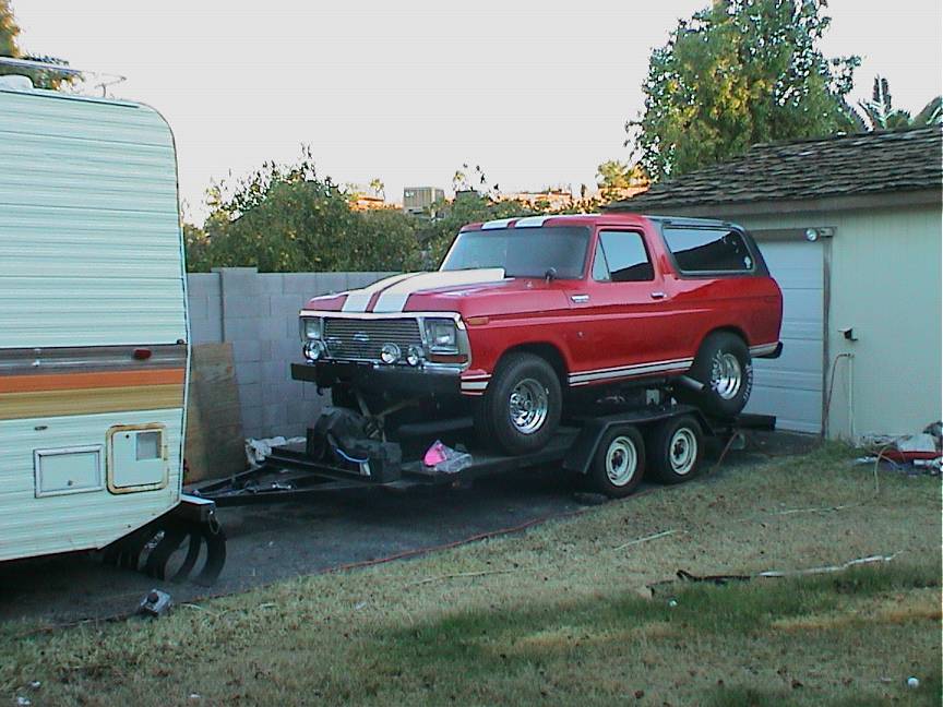 Red Ford Bronco with white hood stripes strapped on a car trailer beside a camper