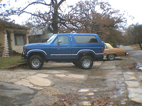 1984 Ford Bronco - A blue SUV is parked on a cracked concrete driveway next to another vehicle.
