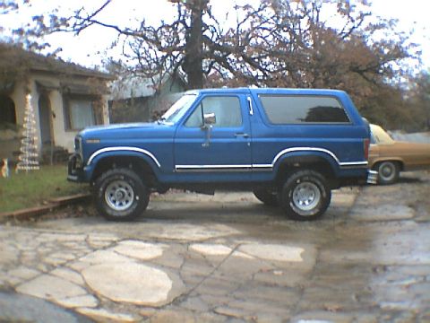 1984 Ford Bronco - A blue SUV is parked on a cracked concrete driveway next to a house and another vehicle.