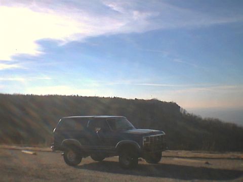 1984 Ford Bronco - A dark SUV is parked on a dirt road overlooking a vast landscape under a cloudy sky.