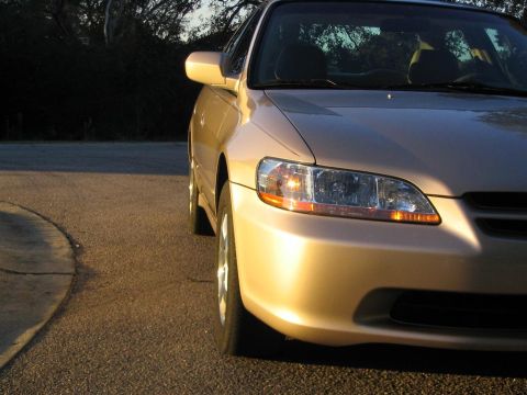 2000 Honda Accord - The front end of a gold sedan is visible, parked on a paved surface with trees in the background.