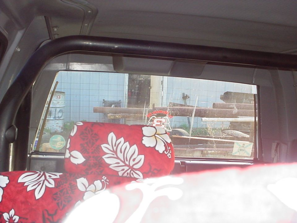 SUV interior with red Hawaiian print seat cover, view out rear window to logs and metal building