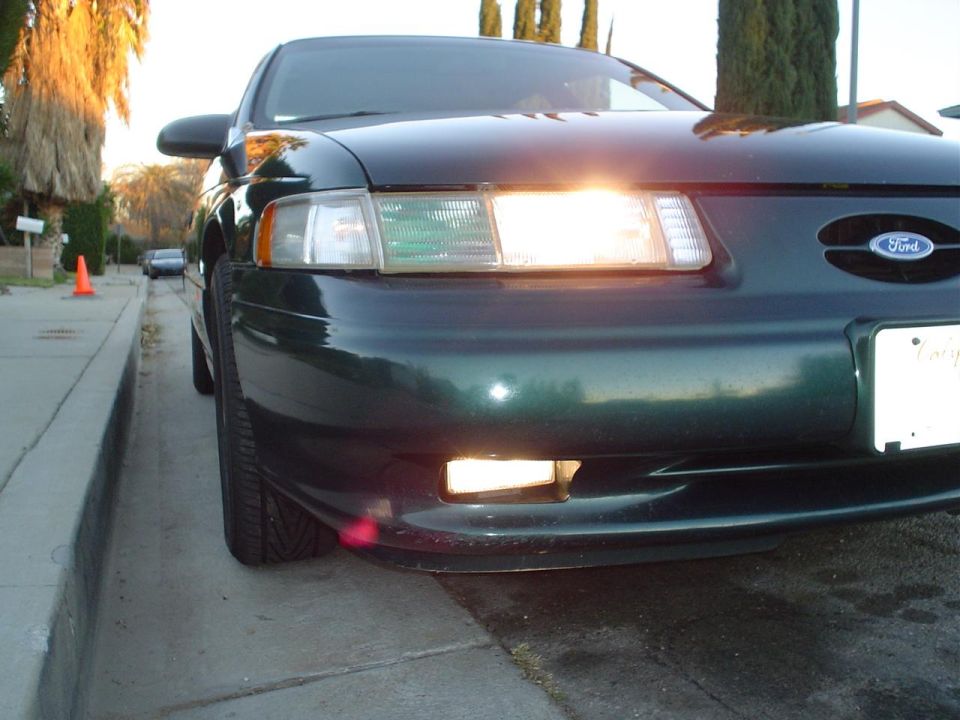 Dark green 1992 Ford Taurus front end, driver's side tire angled towards curb, headlights on