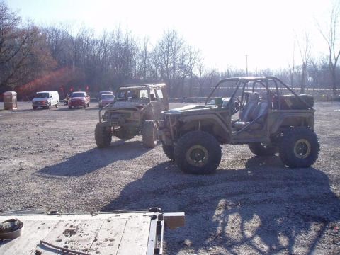 1992 Ford Bronco - Modified off-road vehicles are parked on a gravel lot with trees in the background.