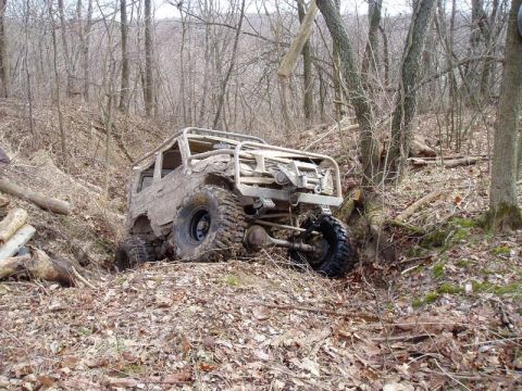 1992 Ford Bronco - This heavily modified off-road vehicle appears stuck in a wooded area, with its front tire deep in a ditch.