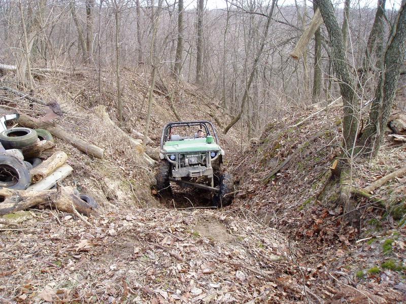 Green off-road vehicle with roll cage navigating a narrow, wooded path with debris on the sides