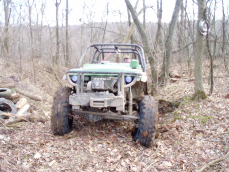 Green off-road vehicle with a roll cage in a wooded area. It has a winch on the front bumper and large tires