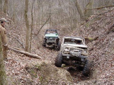 1992 Ford Bronco - Two modified off-road vehicles descend a challenging, muddy path through a dense forest.