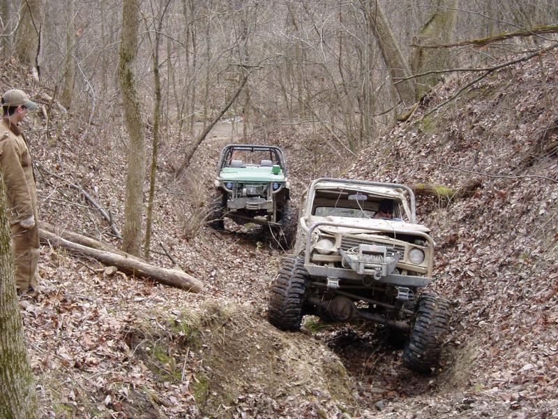Two off-road vehicles navigate a steep, wooded trail; one is tan, the other green