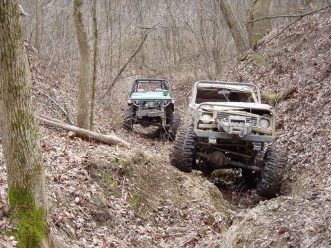 1992 Ford Bronco - Two heavily modified off-road vehicles traverse a challenging trail through a dense forest.