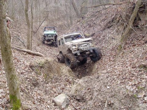 1992 Ford Bronco - Two off-road vehicles tackle a challenging, muddy incline in a wooded area.