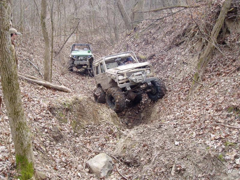 Two off-road vehicles navigating a steep, wooded dirt path. The lead vehicle is white and heavily modified, while the second is green