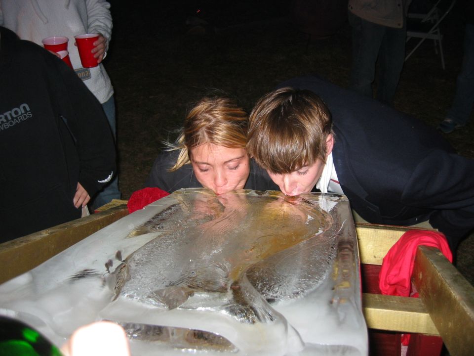 Two people leaning over an ice block luge drinking, partygoers behind them at night