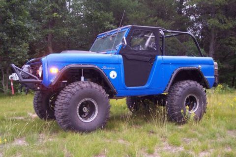 1974 Ford Bronco - Bright blue early Ford Bronco built for off-road, parked in a grassy field with trees behind, seen from the left front three-quarter angle.…