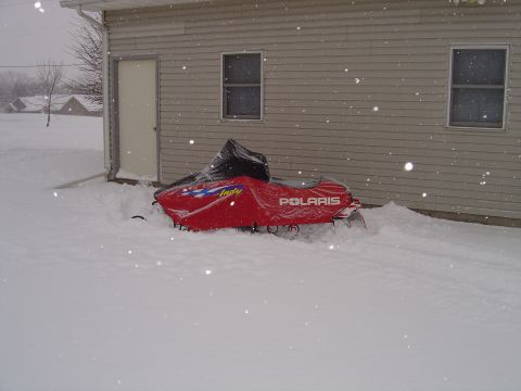 1978 Ford Bronco - A red Polaris snowmobile sits partially buried in snow next to a building during a snowfall.