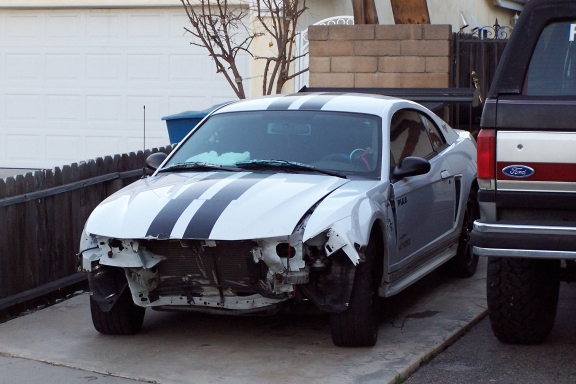 White coupe with black stripes, missing front bumper and grille, parked next to a Ford truck