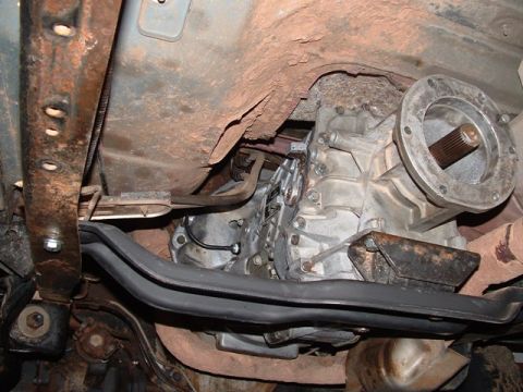 1990 Ford Bronco - Undercarriage shot of an aluminum transfer case with splined output flange, surrounded by a rusty frame and exhaust.