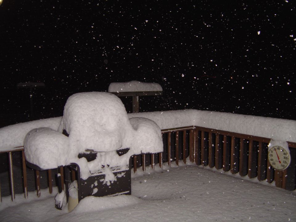 Snow-covered grill on a deck during a snowfall, with a thermometer showing below freezing