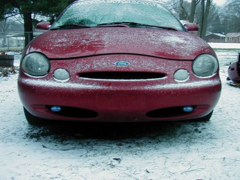 1997 Ford Taurus - A red Ford Taurus sits covered in snow, its front grille and headlights visible.